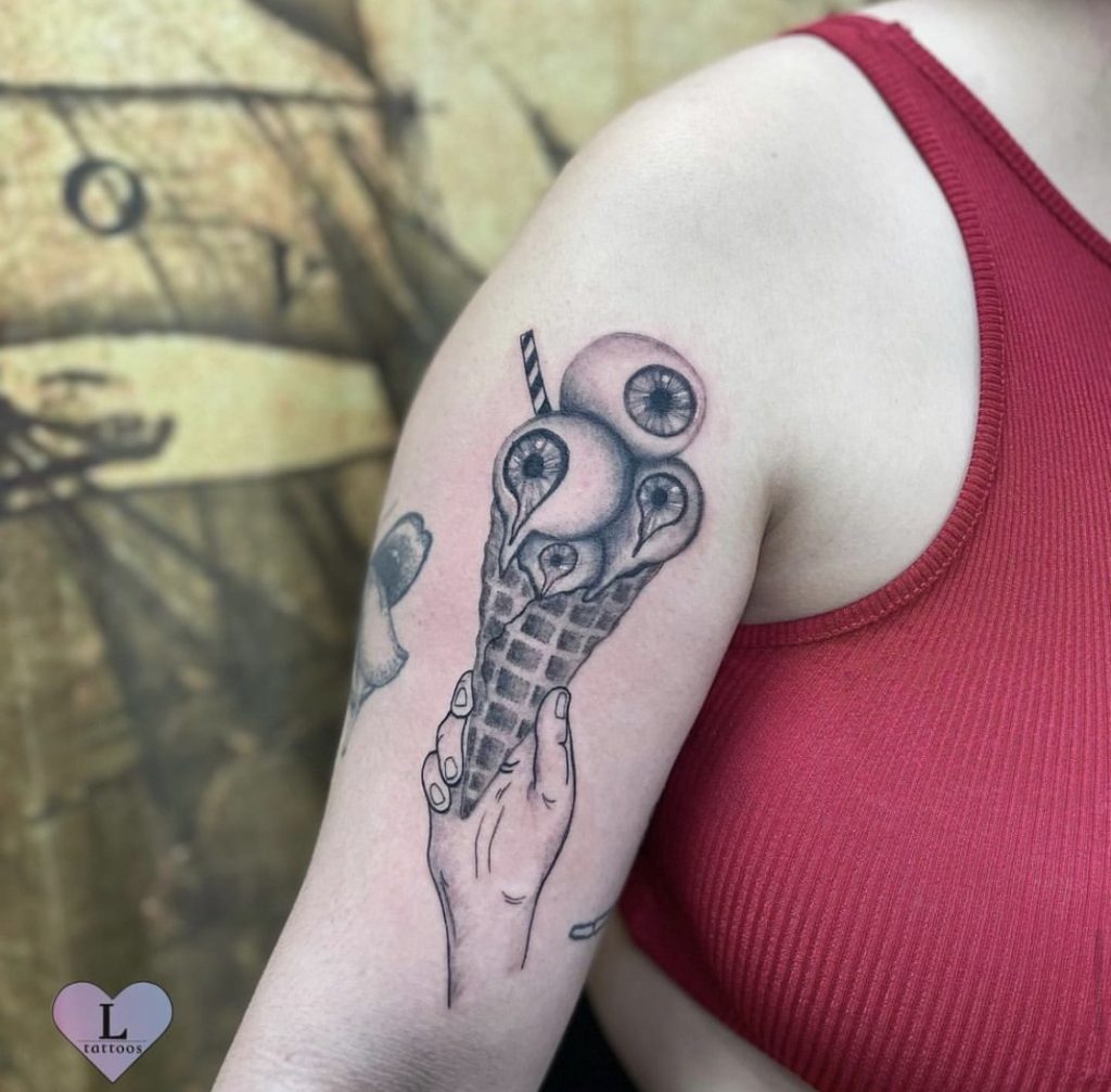 Black and grey eyeball ice cream cone held in a hand, tattooed on an upper arm by Abi Rojas