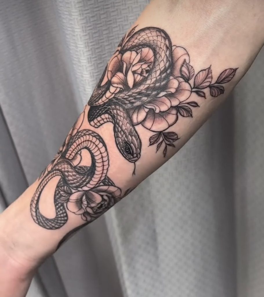 Black and grey snake winding through peonies on a forearm, tattooed by Abi Rojas